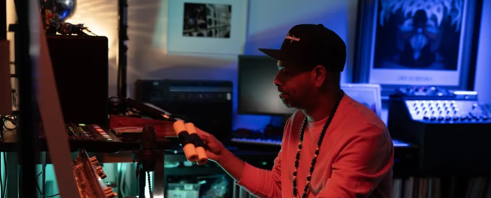 A DJ adjusts equipment in a dimly lit home studio with colorful lighting.