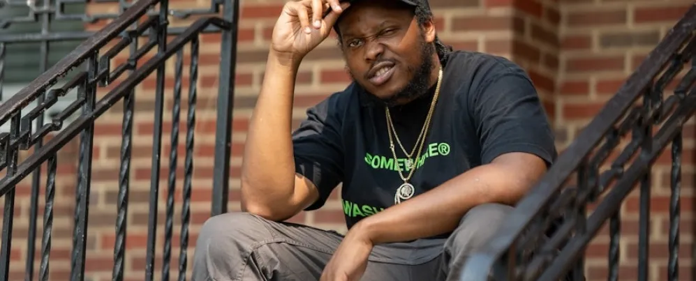 A man in a black T-shirt and cap sits on the steps of a brick building, looking toward the camera.