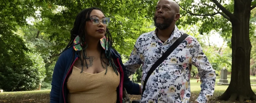 Two people in a leafy park, one holding a phone, both wearing patterned outfits.