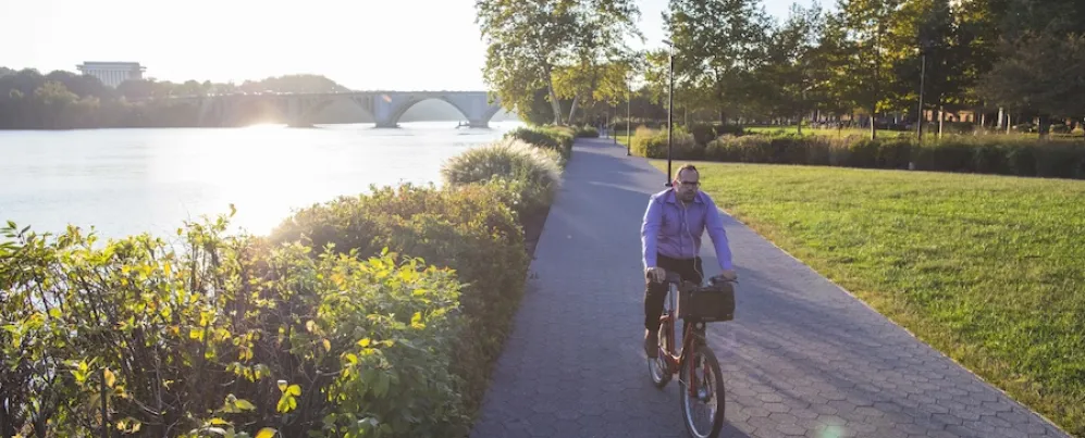 Cyclist on a riverside path at sunset, with trees and green grass nearby