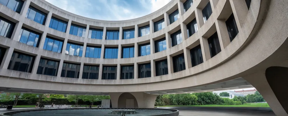 Curved modern building with large windows surrounding a circular fountain under blue sky