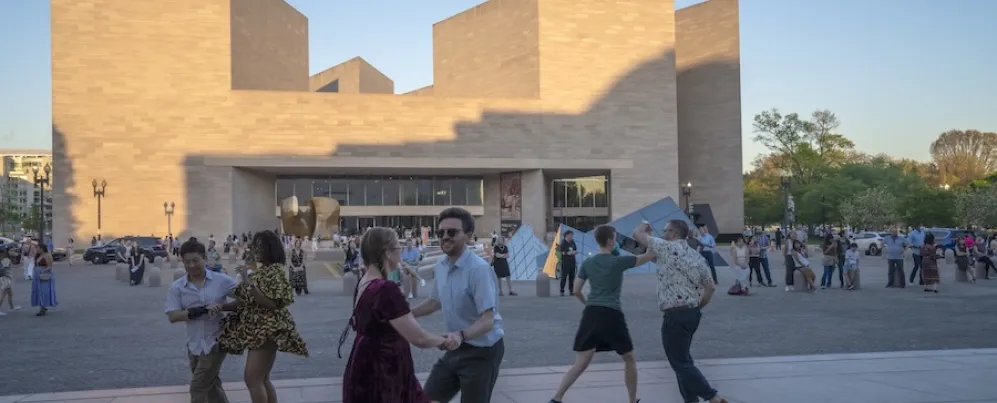 Gente paseando por una plaza frente a un moderno edificio de piedra beige al atardecer.