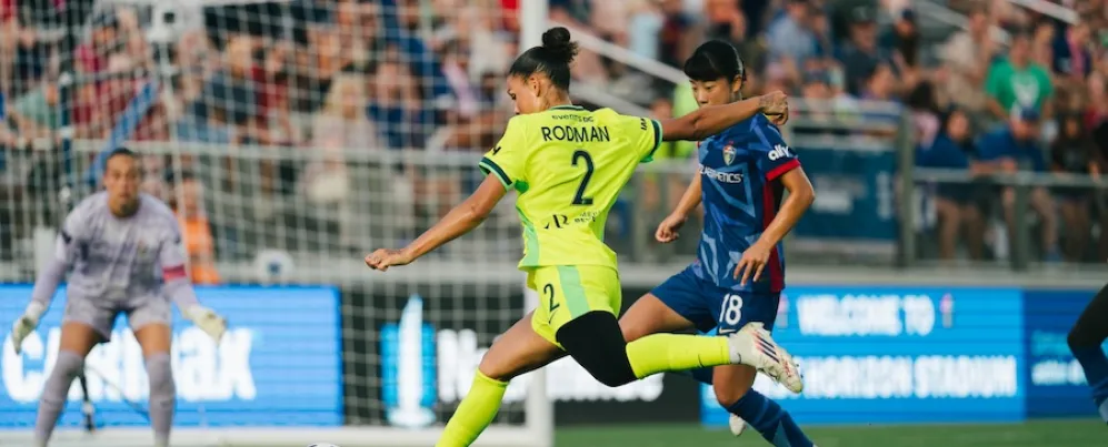 Photo of soccer player in yellow kicking toward goal, defended by blue-uniform player, packed crowd