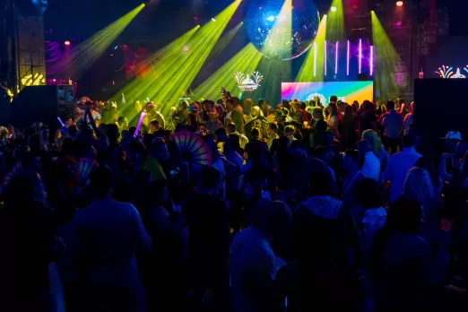 A large, energetic crowd dances under vibrant yellow lights and a disco ball at a colorful Pride celebration event.