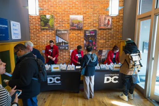 Staff in red jackets assist attendees checking in at a Destination DC event registration table set against a brick wall with Washington, DC promotional posters.
