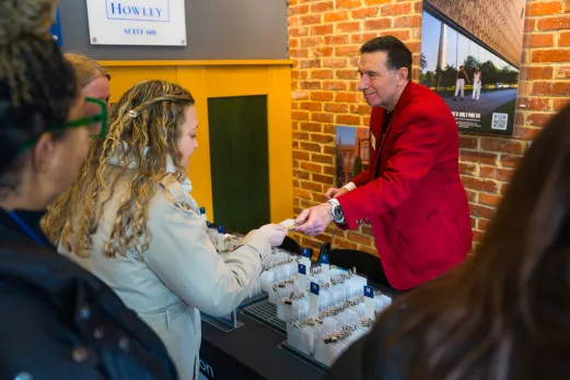 A staff member in a red jacket hands a name badge to an attendee at a Destination DC event check-in table lined with organized badges against a brick wall backdrop.