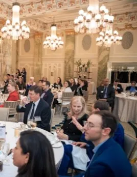 Attendees sit at tables during a formal event in an elegant ballroom illuminated by chandeliers.