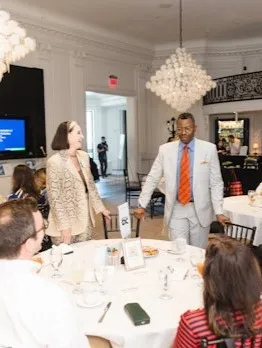 A speaker addresses attendees seated at round tables during an indoor event.