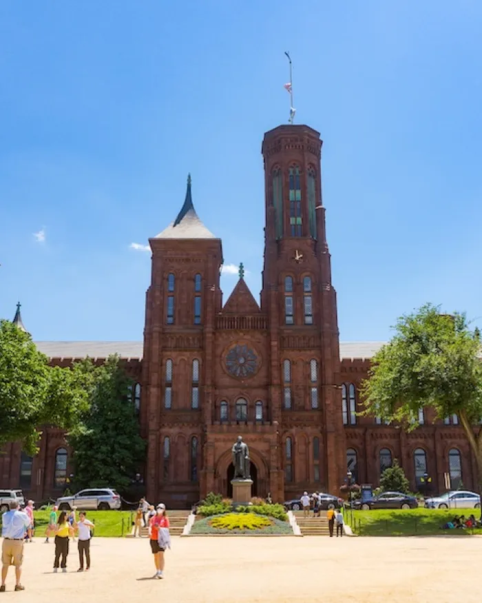 Visitors walk across the National Mall toward the red sandstone Smithsonian Castle on a sunny day.