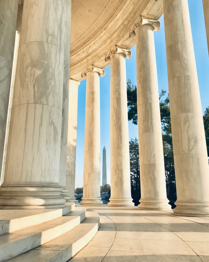 Sunlight filters through the marble columns of the Jefferson Memorial, framing a view of the Washington Monument in the distance.
