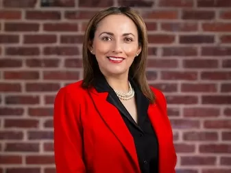 A professional portrait of a woman with shoulder-length hair wearing a red blazer, black shirt, and pearl necklace, smiling in front of a brick wall background.
