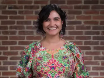 Una persona con i capelli corti e neri, che indossa un vestito con una fantasia colorata, sorride davanti a un muro di mattoni.