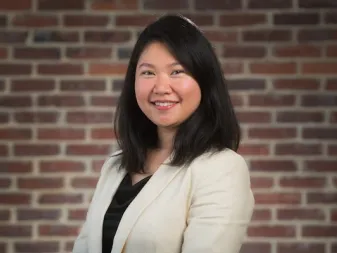 Uma pessoa com cabelos escuros e lisos, vestindo um blazer creme, sorrindo em frente a uma parede de tijolos.