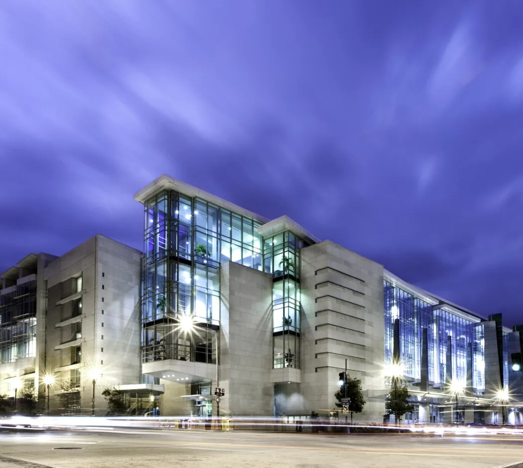 The image shows the exterior of the Walter E. Washington Convention Center in Washington, DC, illuminated at night. The sleek, modern architecture is highlighted by large glass windows and bright lighting, set against a dramatic sky. This convention center is a key venue for conferences, events, and exhibitions in the heart of the nation's capital.