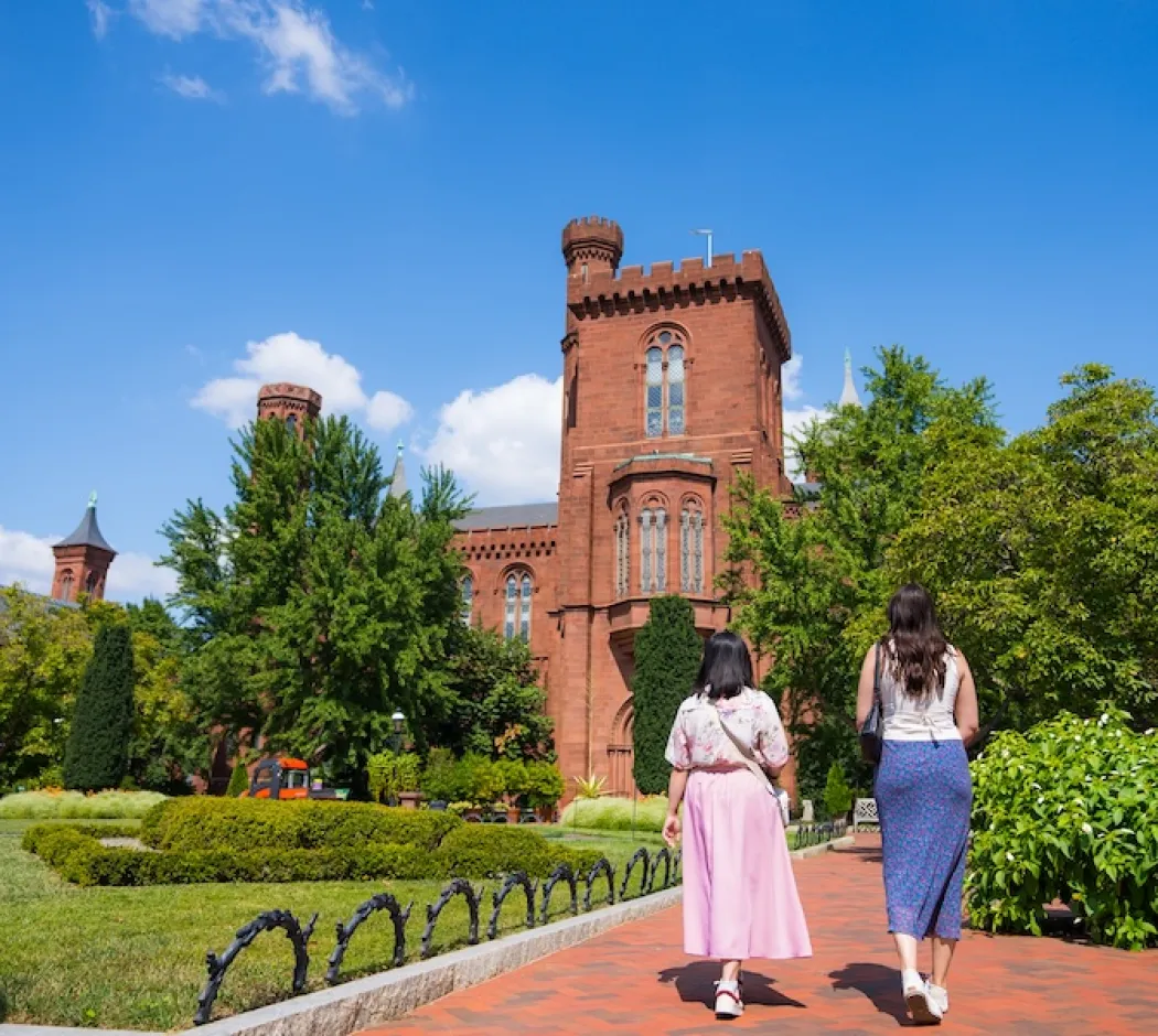 Two women walk along a garden path toward the Smithsonian Castle surrounded by lush greenery.